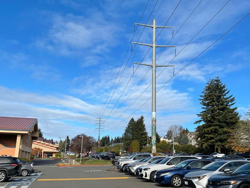 A parking lot with cars in front of a building to the right. There are two grey transmission poles behind the cars with wires running along them.